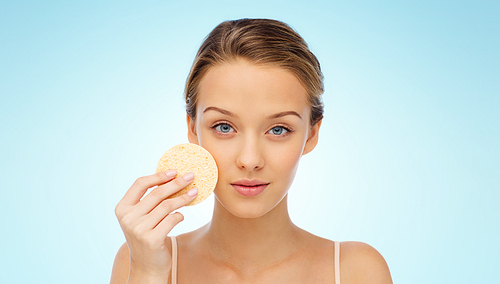 beauty, people and skincare concept - young woman cleaning face with exfoliating sponge over blue background