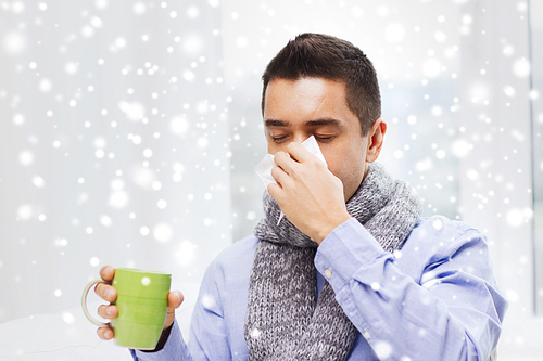 healthcare, flu, people, rhinitis and medicine concept - ill man blowing his nose with paper napkin and drinking tea at home over snow effect
