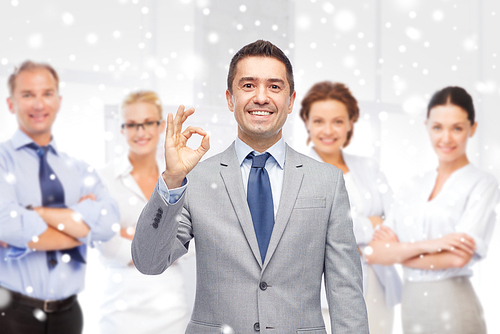 business, people, gesture and success concept - happy smiling businessman in suit with team over office room background showing ok hand sign over snow effect