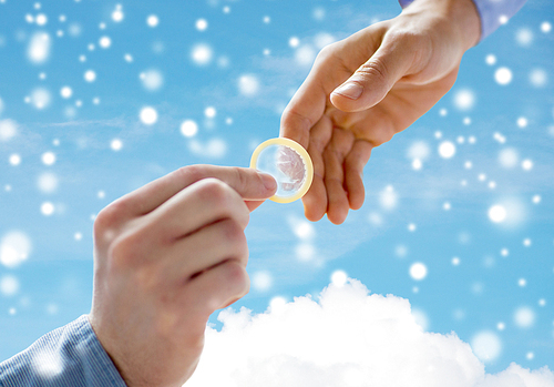 people, homosexuality, safe sex, sexual education and charity concept - close up of happy male gay couple hands giving condom over blue sky and cloud background and snow effect