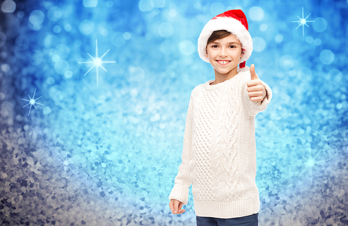 holidays, gesture, christmas, childhood and people concept - smiling happy boy in santa hat showing thumbs up over blue holidays lights background