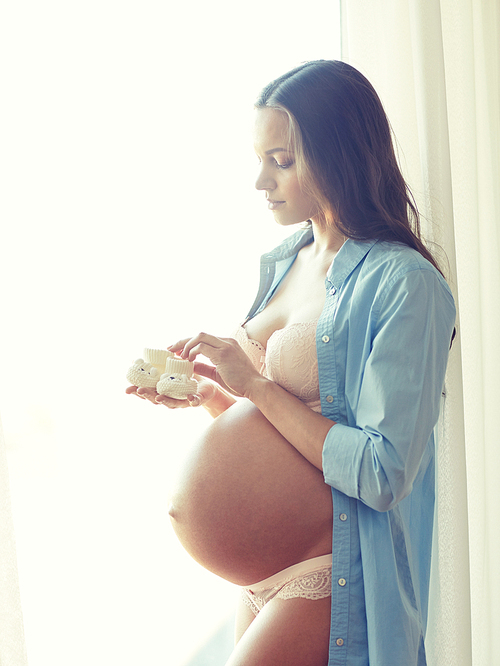 pregnancy, motherhood, people and expectation concept - happy pregnant woman with big bare tummy holding little baby booties at home