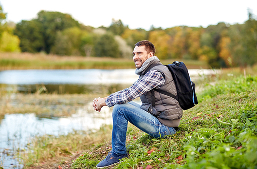 adventure, travel, tourism, hike and people concept - smiling man with backpack resting on river bank