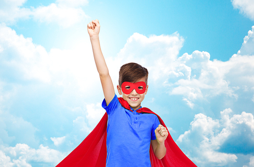 carnival, childhood, power, gesture and people concept - happy boy in red superhero cape and mask showing fists over blue sky and clouds background