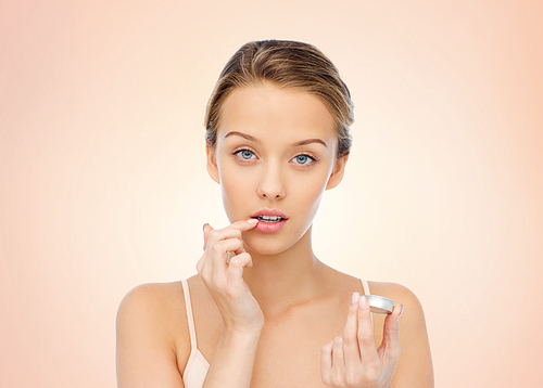 beauty, people and lip care concept - young woman applying lip balm to her lips over beige background