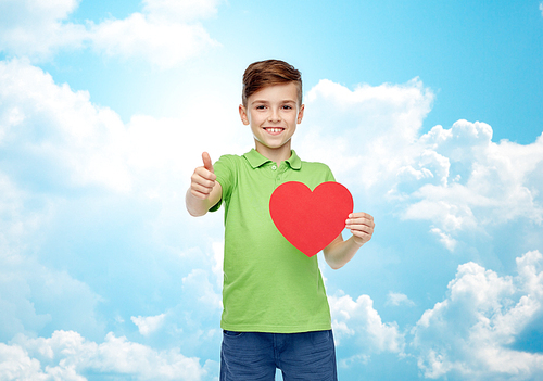 childhood, love, charity, health care and people concept - happy smiling boy in green polo t-shirt holding blank red heart shape and showing thumbs up over blue sky and clouds background