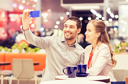 sale, shopping, consumerism, technology and people concept - happy young couple with smartphone taking selfie and drinking coffee or tea at cafe in mall