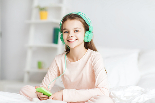 people, children, pajama party and technology concept - happy smiling girl in headphones sitting on bed with smartphone and listening to music at home