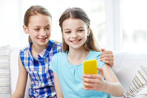 people, children, technology, friends and friendship concept - happy little girls sitting on sofa and taking selfie with smartphone and hugging at home