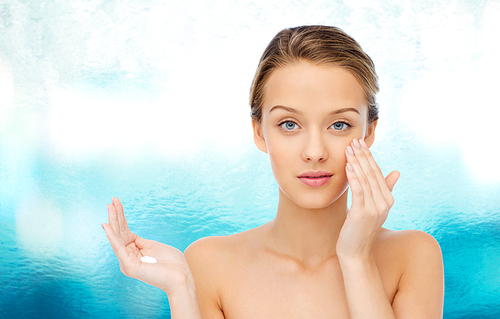 beauty, people, cosmetics, skincare and health concept - young woman applying cream to her face over blue water background