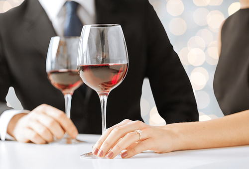 people, holidays, wedding, proposal and jewelry concept - hands of couple with diamond engagement ring and wine glasses in restaurant over lights background
