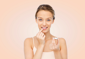 beauty, people and lip care concept - smiling young woman applying lip balm to her lips over beige background