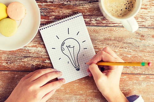 business, education and people concept - close up of female hands with pencil, coffee and cookies drawing lighting bulb to notebook on table