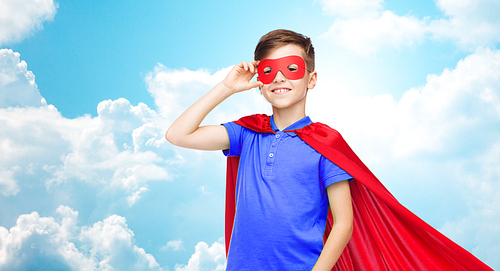 carnival, childhood, power, gesture and people concept - happy boy in red superhero cape and mask over blue sky and clouds background