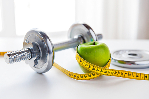 sport, fitness, diet and objects concept - close up of dumbbell and green apple with measuring tape