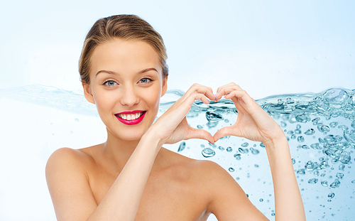 beauty, people, love, valentines day and make up concept - smiling young woman with pink lipstick on lips showing heart shape hand sign over water splash background