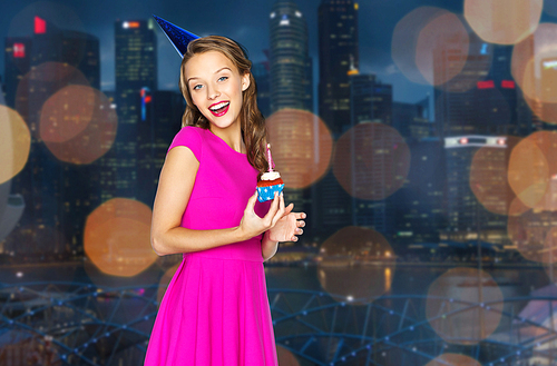 people, holidays and celebration concept - happy young woman or teen girl in pink dress and party cap with birthday cupcake over night singapore city lights background