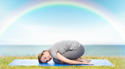 fitness, sport, people and healthy lifestyle concept - happy woman making yoga in child pose on mat over blue sky, rainbow and sea background