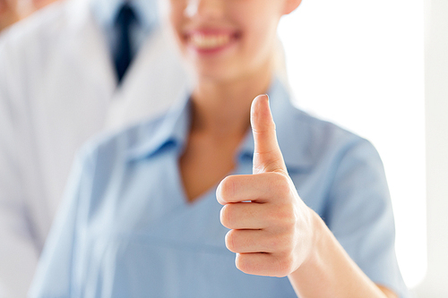 gesture, people, health care and medicine concept - close up of happy female doctor or nurse showing thumbs up hand sign over group of medics meeting at hospital