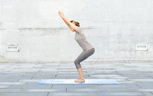 fitness, sport, people and healthy lifestyle concept - woman making yoga in chair pose on mat over urban street background