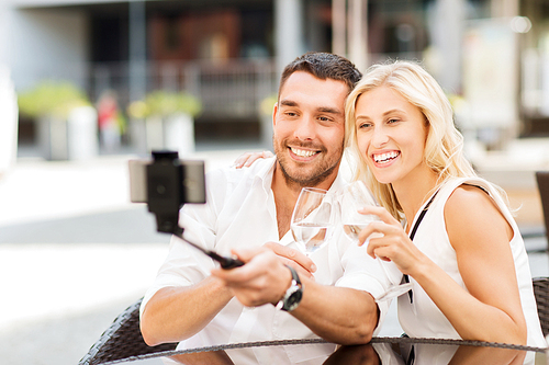 love, date, technology, people and holidays concept - happy happy couple taking picture with smartphone on selfie stick and clinking glasses at city street cafe or restaurant
