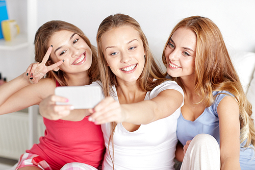 friendship, people, pajama party and technology concept - happy friends or teenage girls with smartphone taking selfie at home