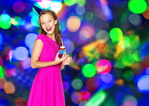 people, holidays and celebration concept - happy young woman or teen girl in pink dress and party cap with birthday cupcake over disco lights background