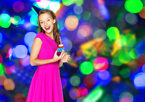 people, holidays and celebration concept - happy young woman or teen girl in pink dress and party cap with birthday cupcake over disco lights background