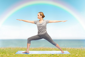 fitness, sport, people and healthy lifestyle concept - woman making yoga warrior pose on mat over blue sky, rainbow and sea background
