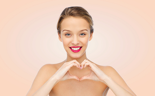 beauty, people, love, valentines day and make up concept - smiling young woman with pink lipstick on lips showing heart shape hand sign over beige background