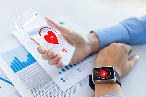 technology, health care and people concept - close up of woman hand holding transparent smartphone and smartwatch with heart icon at office