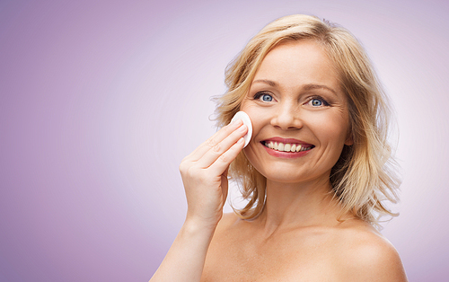 beauty, people and skincare concept - happy middle aged woman cleaning face and removing make up with cotton pad over violet background