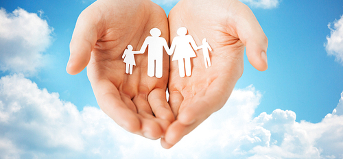 people, values and happiness concept - close up of man cupped hands showing paper family cutout over blue sky and clouds background