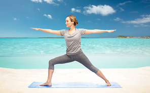 fitness, sport, people and healthy lifestyle concept - woman making yoga warrior pose on mat over sea and sky background