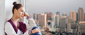 people, emotion and teens concept - sad unhappy pretty teenage girl sitting on windowsill over city background