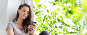 people, technology and teens concept - sad unhappy pretty teenage girl sitting on windowsill with smartphone and texting over summer tree brunch background