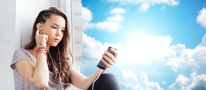 people, technology and teens concept - sad pretty teenage girl sitting on windowsill with smartphone and earphones listening to music over blue sky and clouds background