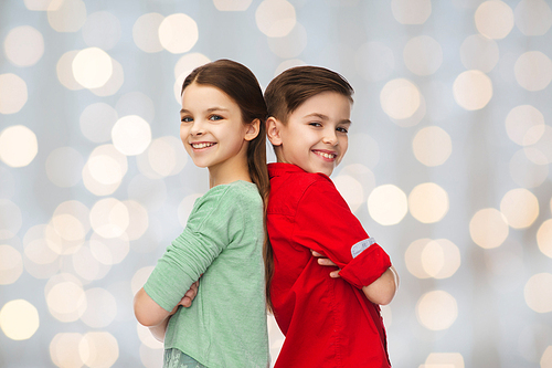 childhood, fashion and people concept - happy smiling boy and girl standing back to back over holidays lights background