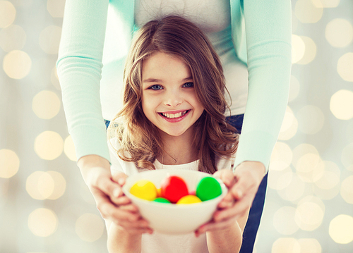 easter, family, people, holiday and childhood concept - close up of happy girl and mother hands holding bowl with colored eggs over lights background