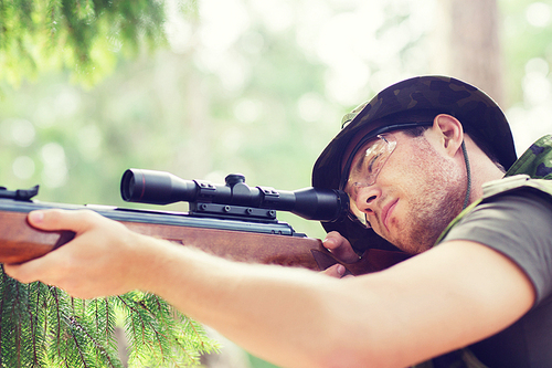 hunting, war, army and people concept - young soldier, ranger or hunter with gun aiming and shooting in forest