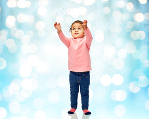 childhood, fashion, clothing and people concept - beautiful little african american baby girl playing with soap bubble over blue holidays lights background