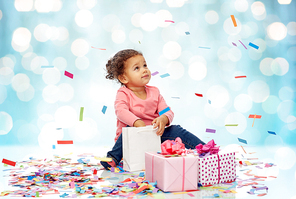childhood, birthday, party, holidays and people concept - happy little african american baby girl with gift boxes and confetti playing with shopping bag sitting on floor over blue lights background