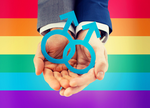 people, homosexuality, same-sex marriage and love concept - close up of happy male gay couple holding paper cutout love symbol over rainbow flag background