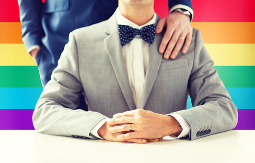 people, celebration, homosexuality, same-sex marriage and love concept - close up of male gay couple with wedding rings on putting hand on shoulder over rainbow flag background