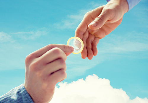 people, homosexuality, safe sex, sexual education and charity concept - close up of happy male gay couple hands giving condom over blue sky and cloud background