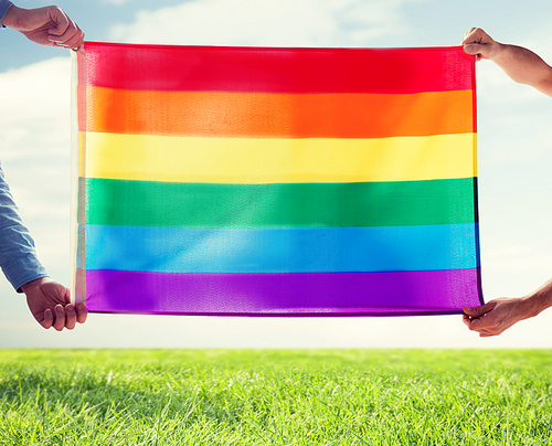 people, homosexuality, same-sex, gay and love concept - close up of happy male gay couple hugging and holding rainbow flag over blue sky and grass background