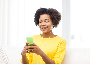 people, technology and leisure concept - happy african american young woman sitting on sofa with smartphone and earphones listening to music at home