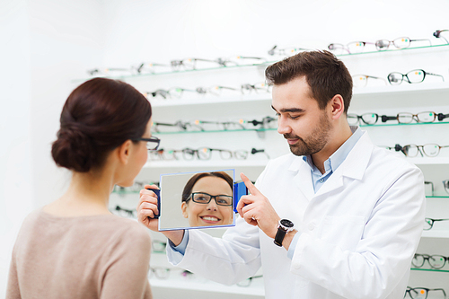 health care, people, eyesight and vision concept - optician and woman in glasses looking to mirror at optics store