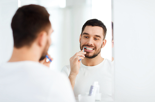 health care, dental hygiene, people and beauty concept - smiling young man with toothbrush cleaning teeth and looking to mirror at home bathroom