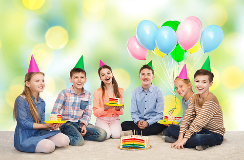 childhood, holidays, celebration, friendship and people concept - happy smiling children in party hats with birthday cake and balloons over green summer holidays lights background